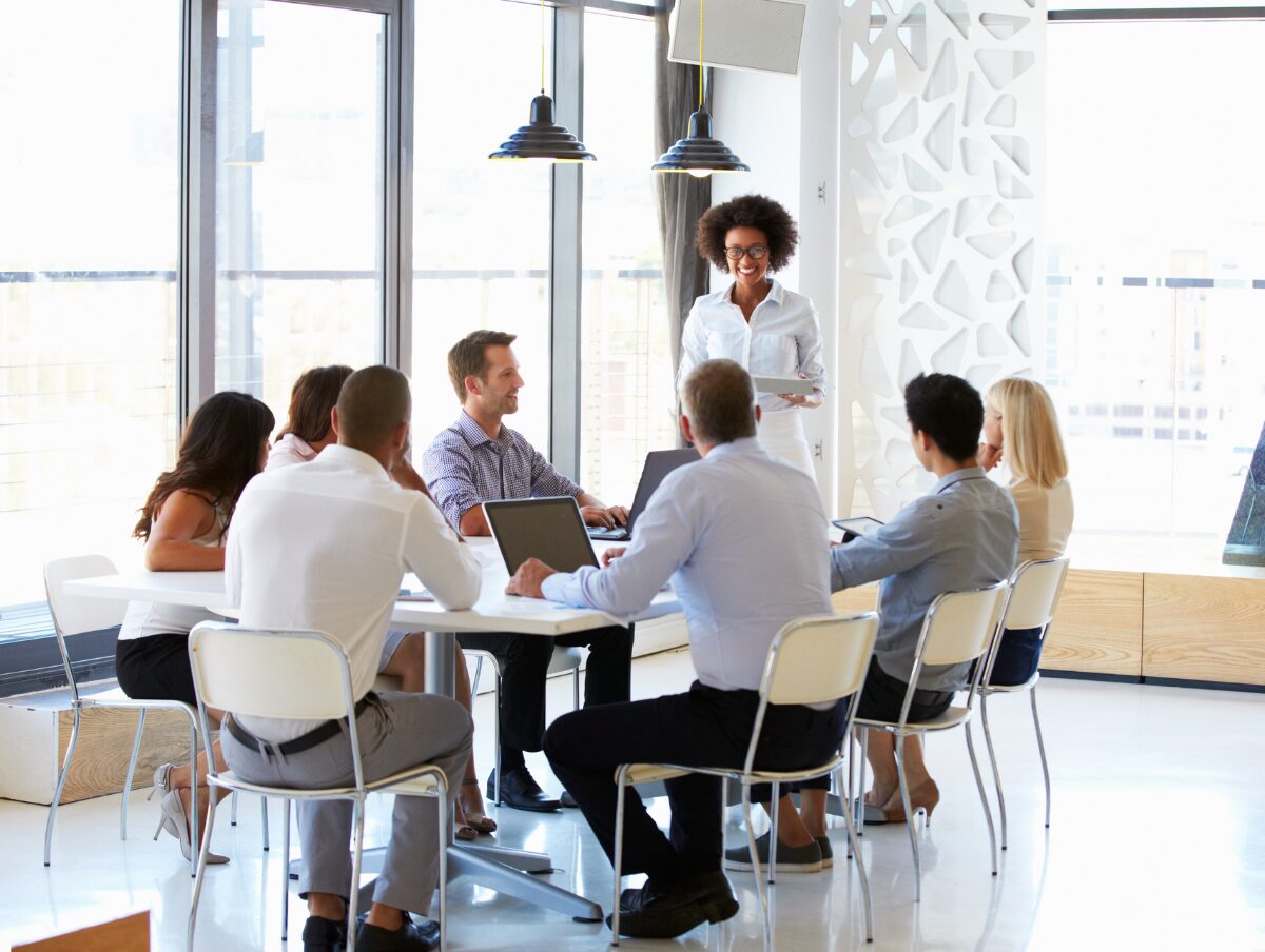 Un groupe de personnes dans une salle de conférence assises autour d'une table tandis que quelqu'un est debout et parle, avec de grandes fenêtres en arrière-plan. Un groupe de personnes dans une salle de conférence assises autour d'une table tandis que quelqu'un est debout et parle, avec de grandes fenêtres en arrière-plan.