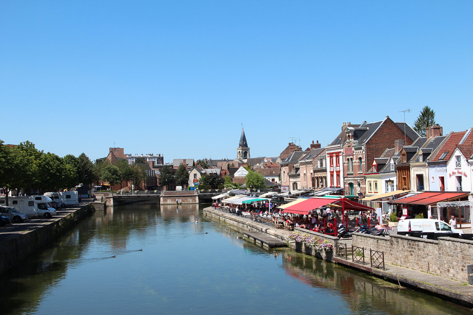 Une rivière calme bordée de bâtiments colorés et de cafés en plein air sous un ciel bleu clair, avec un clocher d'église visible en arrière-plan. Une rivière calme bordée de bâtiments colorés et de cafés en plein air sous un ciel bleu clair, avec un clocher d'église visible en arrière-plan.
