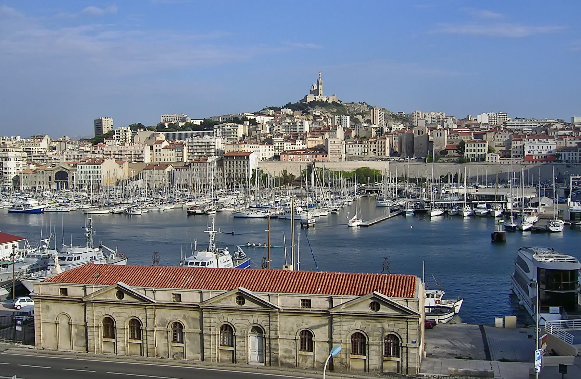 Vue d'une marina avec de nombreux bateaux amarrés, des bâtiments bordant le front de mer et une église au sommet d'une colline en arrière-plan sous un ciel bleu clair. Vue d'une marina avec de nombreux bateaux amarrés, des bâtiments bordant le front de mer et une église au sommet d'une colline en arrière-plan sous un ciel bleu clair.